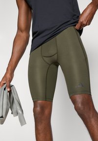 Pantalones cortos ajustados de color verde oliva con una textura suave, que tienen una longitud hasta medio muslo, un bolsillo con costura y un logo negro en el lateral.