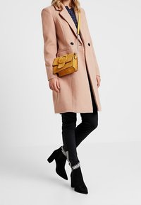 Cappotto rosa pallido con bottoni neri, borsa a tracolla gialla con dettaglio a fiocco, pantaloni neri con strisce bianche e stivaletti neri con tacco.