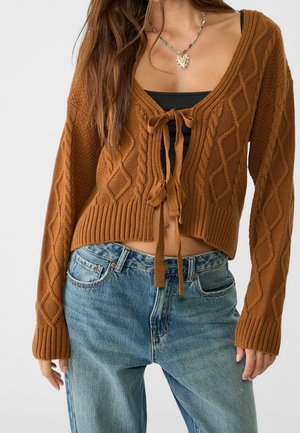 Femme portant un cardigan court en maille torsadée marron noué à l'avant sur un haut noir et un jean bleu taille haute, avec un collier en forme de cœur.