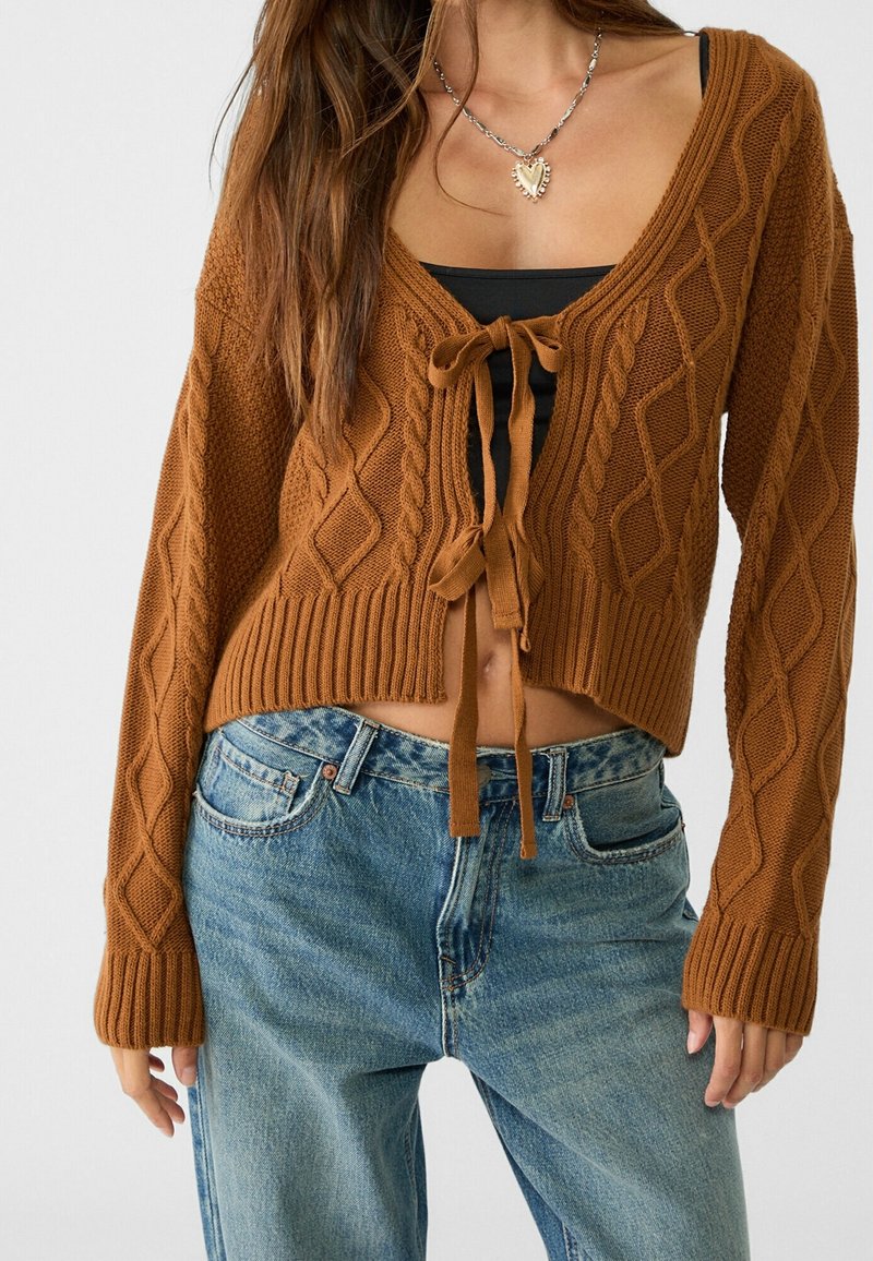 Femme portant un cardigan court en maille torsadée marron noué à l'avant sur un haut noir et un jean bleu taille haute, avec un collier en forme de cœur.