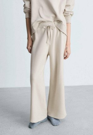 Personne portant un pantalon beige à jambes larges et un sweat assorti, debout pieds nus sur un sol gris clair avec un fond uni.