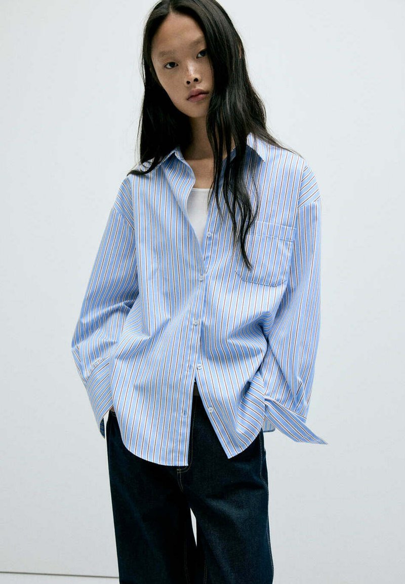 Jeune femme aux longs cheveux noirs portant une chemise oversize rayée bleu et blanc par-dessus un top blanc et un pantalon sombre, sur un fond uni.