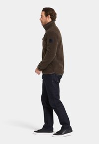 Bruine fleece pullover met een hoge kraag, textuur op het oppervlak en een patch op de linkerarm, gecombineerd met donkere denim jeans en zwarte sneakers.