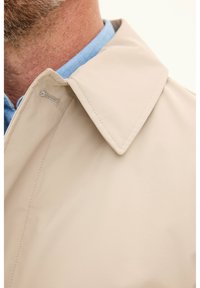 Beige outerwear with a structured collar, featuring smooth fabric and a visible seam detail on the collar. Worn over a light blue shirt.