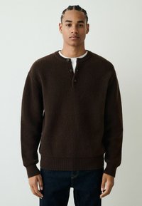 Brown ribbed knit sweater with a round neck and two-button placket, worn over a white shirt. Features long sleeves and a relaxed fit.