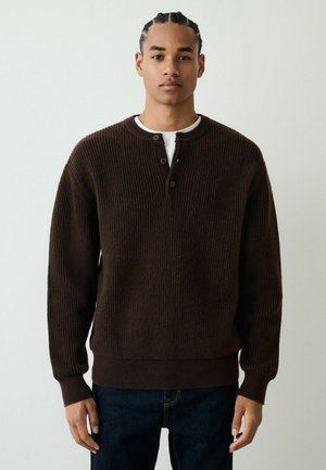 Brown ribbed knit sweater with a round neck and two-button placket, worn over a white shirt. Features long sleeves and a relaxed fit.