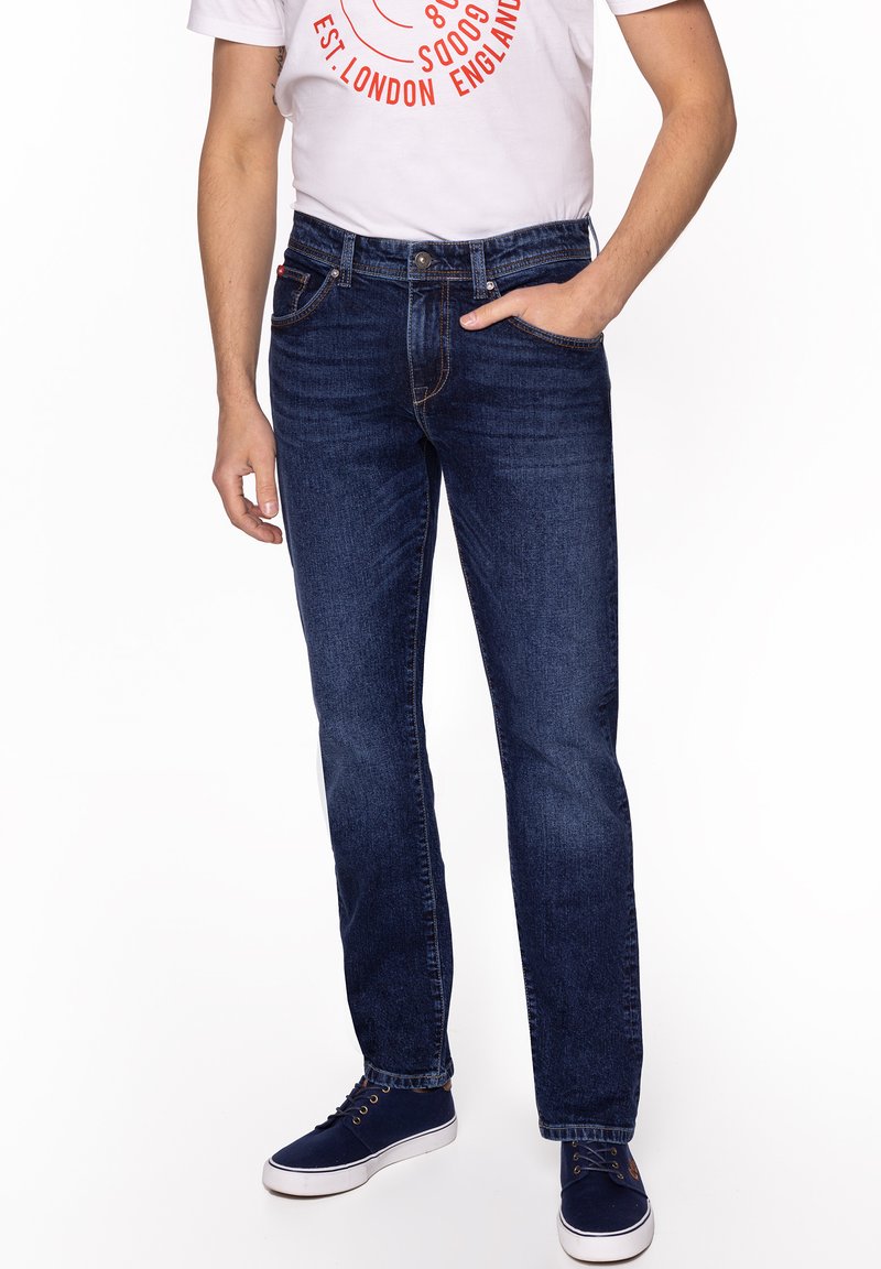 Mężczyzna w ciemnoniebieskich dżinsach, granatowych tenisówkach i białej koszulce z czerwonym napisem, stojący z jedną ręką w kieszeni.