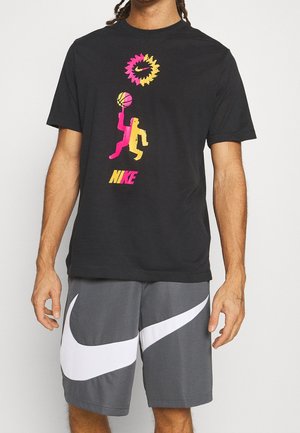 Mann trägt schwarzes Nike-T-Shirt mit buntem Basketballspieler-Grafik und graue Nike-Shorts mit großem weißem Swoosh-Logo.