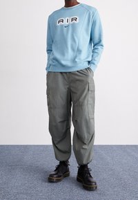 Ljusblå sweatshirt med "AIR" grafik och Nike-logotyp, i kombination med gröna cargobyxor och svarta snörade platåstövlar. Slät textur, avslappnad passform.
