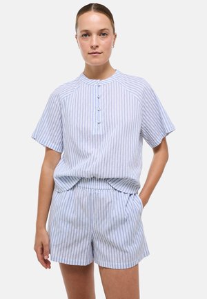 Femme portant un haut boutonné à manches courtes rayé verticalement bleu clair et blanc, et un short assorti à taille élastique, debout avec une main dans la poche.