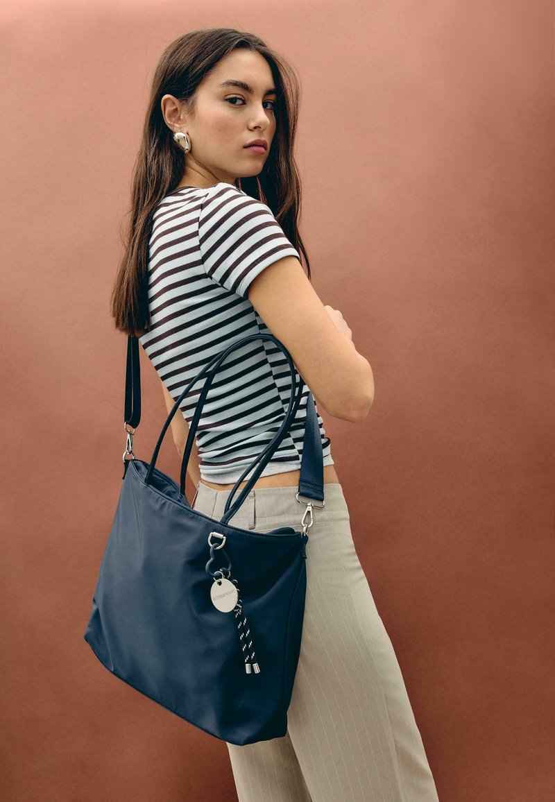 Jeune femme aux longs cheveux bruns, portant une chemise à rayures et un pantalon beige, tenant un grand sac à main bleu marine sur un fond marron uni.