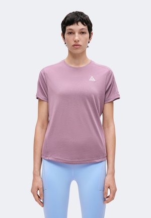 Personne aux cheveux courts et foncés portant un t-shirt violet à manches courtes et des leggings bleu clair, debout devant un fond uni.