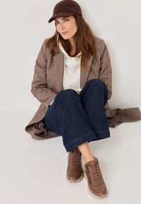 Hnědý tweedový kabát přes bílou mikinu, v kombinaci s tmavými denimovými kalhotami a hnědými teniskami. Čepice je v odpovídající tmavé barvě.