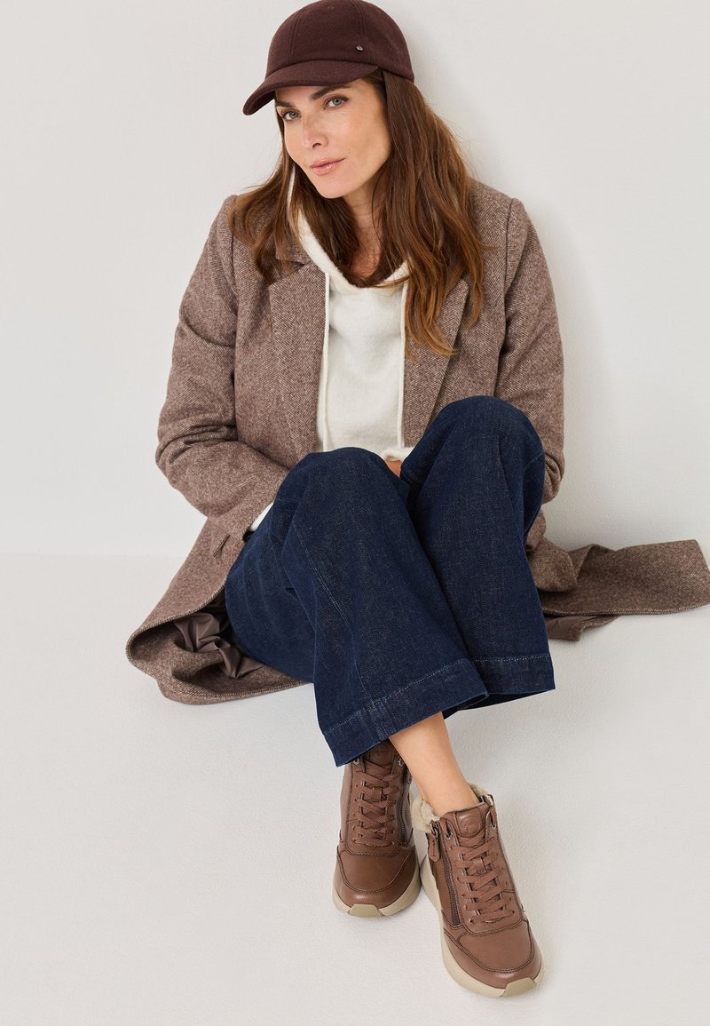 Hnědý tweedový kabát přes bílou mikinu, v kombinaci s tmavými denimovými kalhotami a hnědými teniskami. Čepice je v odpovídající tmavé barvě.