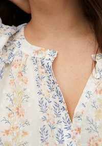 Biała bluzka w niebieskie i brzoskwiniowe wzory kwiatowe, częściowo zapięta na kołnierzyku, noszona przez osobę z długimi brązowymi włosami.