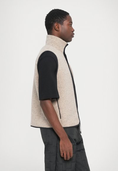 Beige Fleece-Weste mit hohem Kragen, schwarzem Reißverschluss und kontrastierenden schwarzen Ärmeln. Über einem schwarzen kurzärmeligen Hemd und schwarzen Hosen getragen.
