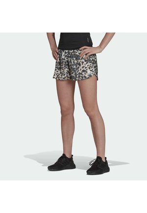 Person trägt schwarze Sneakers und Adidas-Laufshorts mit Leopardenmuster, die Hände in die Hüften gestützt, vor schlichtem Hintergrund.