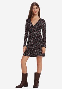 Svart blommig klänning med långa ärmar, V-hals och åtsittande midja, prydd med små röda och vita blommor. Bärs med bruna knähöga stövlar.