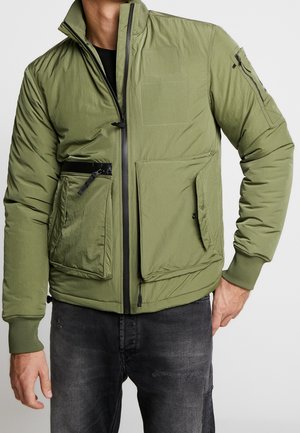 Mann trägt olivgrüne Bomberjacke mit mehreren Reißverschluss- und Klappentaschen, kombiniert mit ausgewaschenen schwarzen Jeans, steht mit einer Hand in der Tasche.