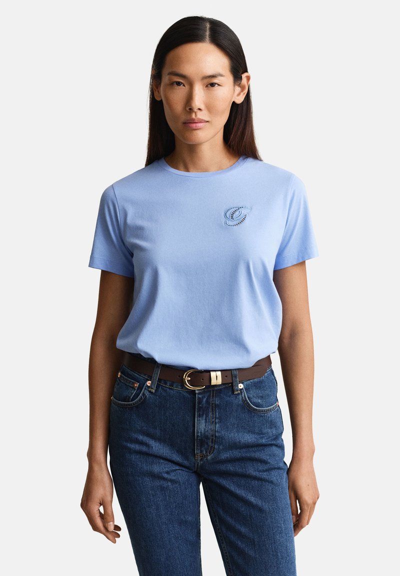 Femme aux longs cheveux bruns portant une chemise bleu clair à manches courtes avec un petit logo brodé et un jean bleu taille haute avec une ceinture marron.