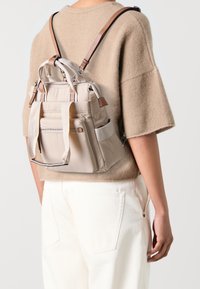 Persona con un suéter beige de manga corta y pantalones blancos, llevando en la espalda una pequeña mochila de tela beige con correas de cuero.