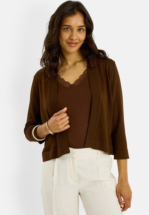 Femme aux cheveux bruns ondulés portant un cardigan marron, un haut avec bordure en dentelle, un pantalon blanc et des bracelets dorés sur un fond clair.