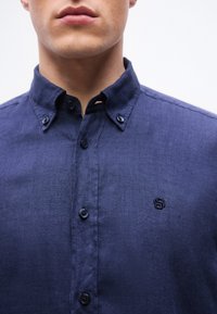 Άνδρας που φοράει πουκάμισο από λινό σε ναυτικό μπλε χρώμα, με κουμπιά και γιακά με κουμπί και μικρό κεντητό λογότυπο στο στήθος.