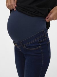 Blå graviditetsjeans med elastisk marineblå brystpanel, tettsittende design og fem-lommers styling. Stoffet ser ut som denim med lett glans.