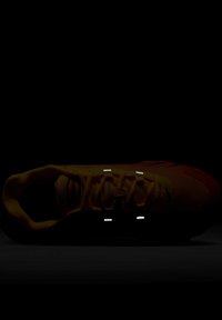 Sárga sportcipő struktúrált felsőrésszel, kontrasztos piros díszítésekkel, fényvisszaverő részletekkel, párnázott bokarésszel és lapos fűzőkkel.