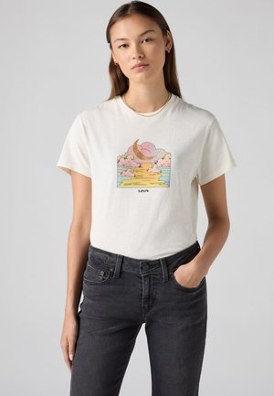T-shirt di cotone color crema con un grafico di una luna e nuvole in tonalità pastello, abbinata a jeans grigio scuro.
