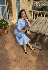 Žena u svijetloplavoj haljini i crnim sandalama sjedi na drvenoj klupi na popločanom trijemu uz saksiju sa žutim cvijećem.