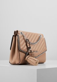 Tan vävd läderhandväska med silverfärgad kedjeslinga, pärlprydnad och två matchande tofscharms som vilar på en vit yta.