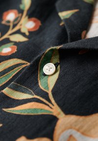 Tessuto scuro con un motivo floreale in arancione, verde e crema. Un bottone crema è visibile, etichettato "SUPERB". Tessitura di lino texturizzato.