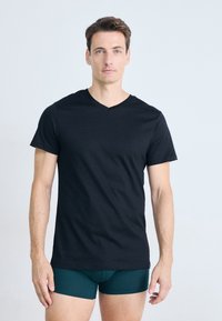 Homme avec des cheveux bruns courts portant un t-shirt noir à col en V et un boxer vert foncé, se tenant contre un fond clair plat.