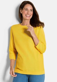 Gelber Strickpullover mit rundem Ausschnitt, Dreiviertelärmeln und geripptem Saum; getragen mit blauen Jeans. Glatte Textur.