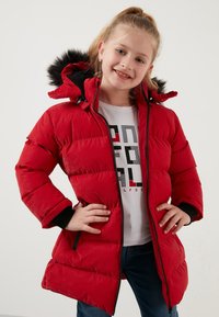 Rød pufferjakke med sorte accenter, aftagelig hætte og faux fur kant. Har en fuld lynlås foran og ribbede manchetter, båret over en hvid t-shirt.