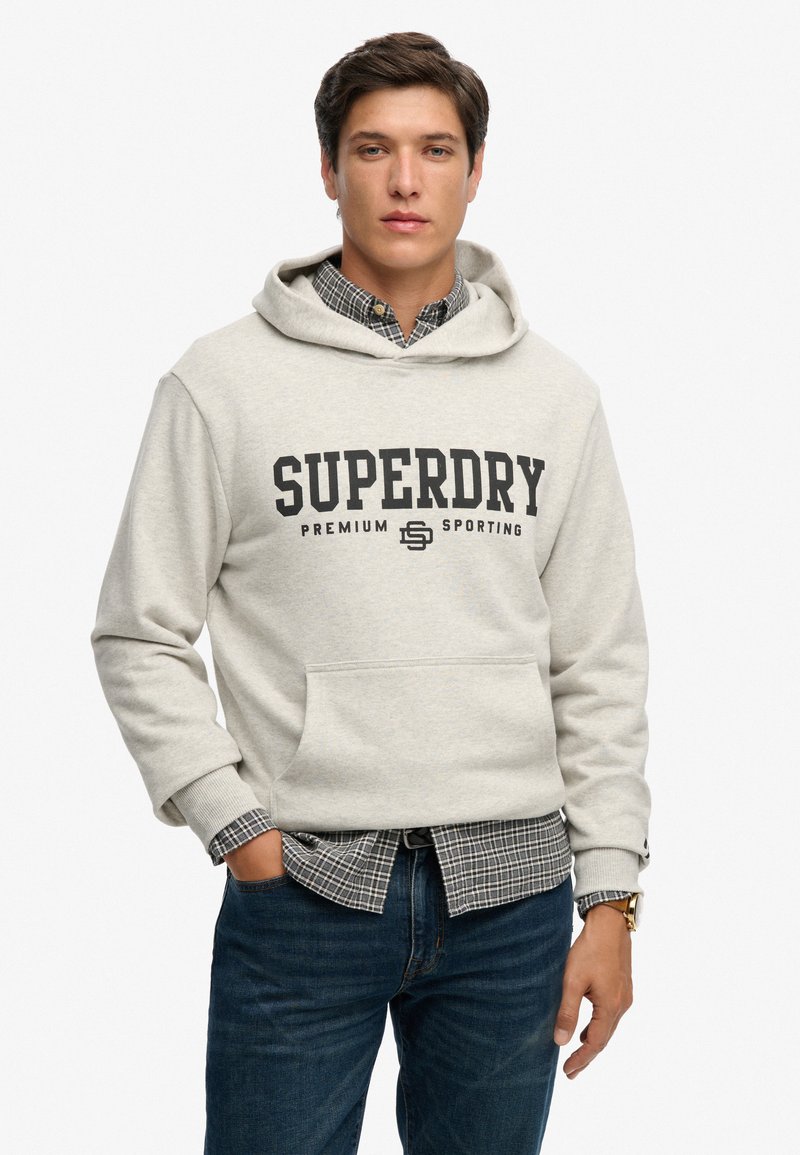 Grijze hoodie met "SUPERDRY" in het zwart gedrukt. Voorzien van een voorzak, een capuchon met trekkoorden en gestructureerde manchetten. Gestyled over een geruit overhemd.