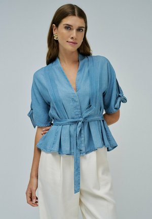 Modello che indossa un top in denim azzurro chiaro con design a peplum e vita cinturata. Ha lunghi capelli castani e eleganti orecchini dorati.