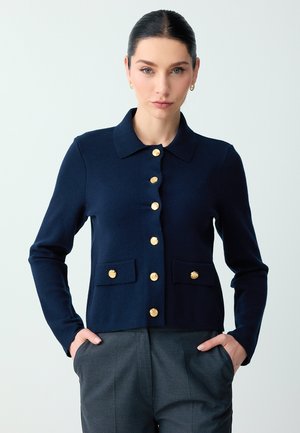 Frau mit dunklem Haar, die eine marineblaue, geknöpfte Strickjacke mit goldenen Knöpfen und graue Hosen trägt, steht mit den Händen in den Taschen.