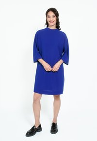 Modra kratkosleeve obleka ohlapnega kroja in visokim ovratnikom. V kombinaciji s črnimi platform škornji. Gladko blago, brez vidnih vzorcev ali dodatkov.