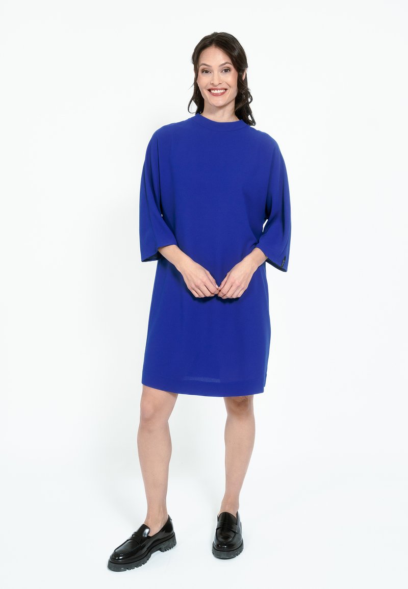 Modra kratkosleeve obleka ohlapnega kroja in visokim ovratnikom. V kombinaciji s črnimi platform škornji. Gladko blago, brez vidnih vzorcev ali dodatkov.