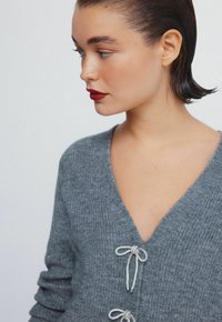 Szary sweter dzianinowy z dekoltem w kształcie litery V, z ozdobnymi srebrnymi wstążkami z przodu. Miękka tekstura i szczegóły w żebrowanym wzorze.