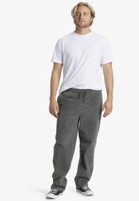 Hombre con cabello rubio, vestido con una camiseta blanca, pantalones grises holgados con cordón y zapatillas blanco y negro, de pie sobre un fondo blanco.