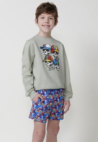 Sudadera de felpa verde claro con un gráfico de calaveras con gorras de colores. Combinada con pantalones cortos azules que exhiben patrones vibrantes de calaveras y tibias cruzadas.