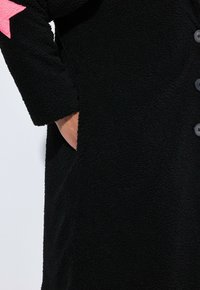 Cappotto nero con texture, bottoni, una mano nella tasca laterale e un dettaglio a stella rosa sulla manica superiore.