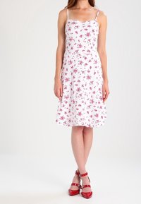 Bílá květinová šaty s tenkými ramínky, které mají malé růžové květy, rozšířenou sukni a lodičkový výstřih. Doplňují je červené sandály na řemínky.
