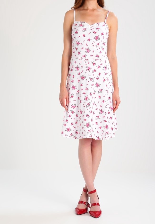 Bílá květinová šaty s tenkými ramínky, které mají malé růžové květy, rozšířenou sukni a lodičkový výstřih. Doplňují je červené sandály na řemínky.