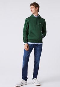 Jeune homme souriant et regardant sur le côté, portant un sweat à capuche vert par-dessus une chemise bleue, un jean bleu et des baskets blanches, debout sur un fond uni.