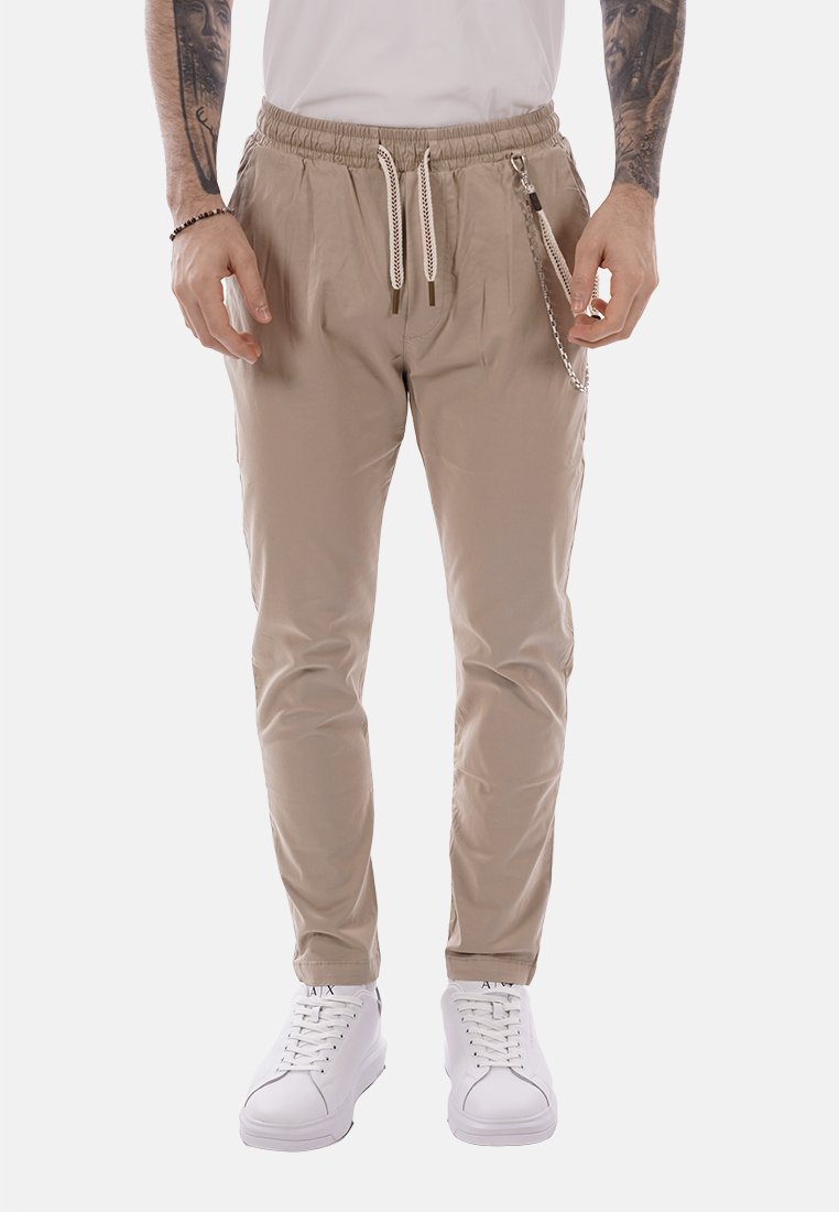 Uomo che indossa pantaloni beige con coulisse e accessorio a catena, e sneakers bianche, in piedi davanti a uno sfondo bianco.