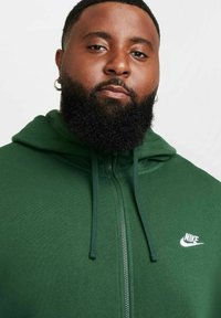 Grön zip-up hoodie i mjukt tyg, med framdragkedja, dragskohuva och en vit Nike-logotyp på vänster bröst.